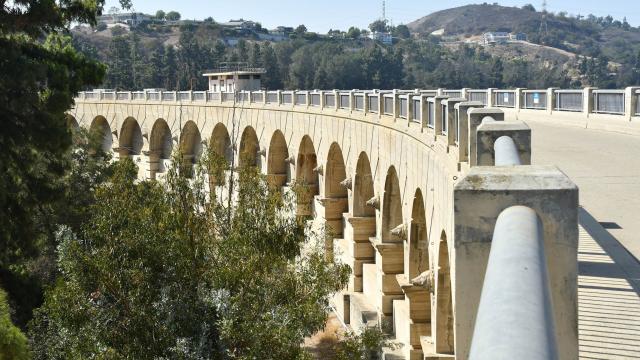 Mulholland Dam showing its curved concrete details surrounded by foliage on the left with hillsides visible in the background.