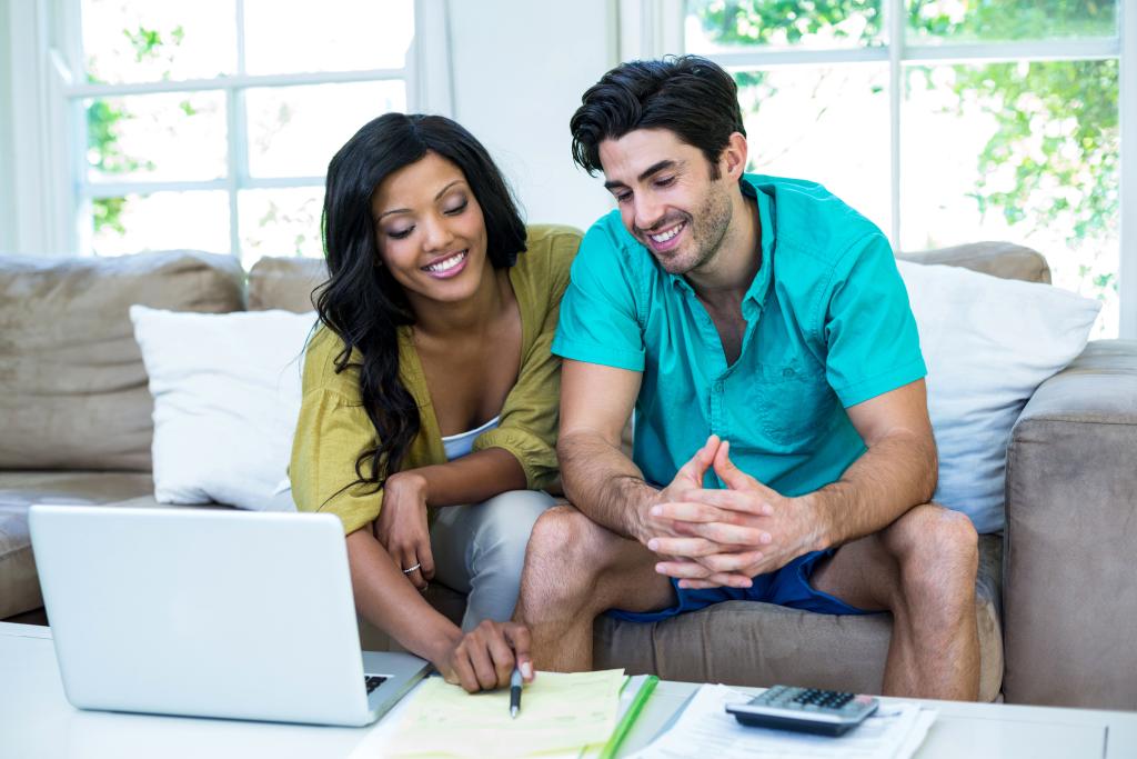 Smiling couple sitting on a couch in front of a table that has a laptop, notepad, and calculator. The woman is holding a pen and pointing to the paper and the man is looking at the paper.