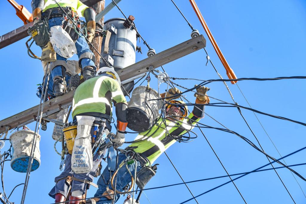 Three lineworkers in hardhats and yellow, reflective safety vests perched atop a power pole as they conduct repairs on electrical lines during a heat storm.