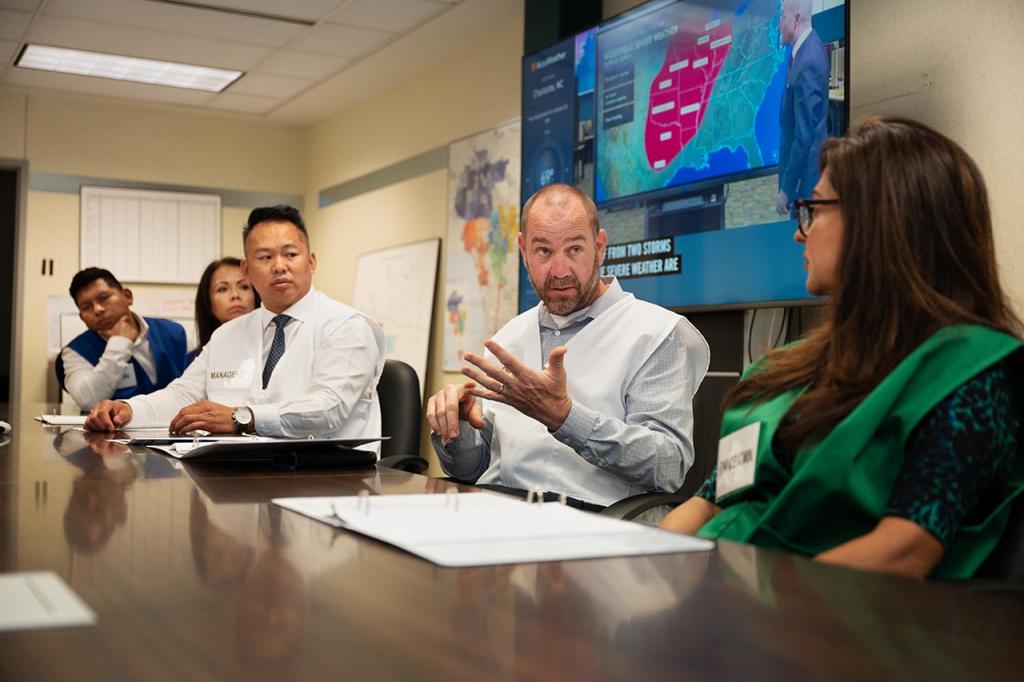 Three men and two women sitting at a conference room table. The man in the center is explaining something using his fingers to count off. Behind them are several maps, charts and a flat screen television showing a weathercast.  