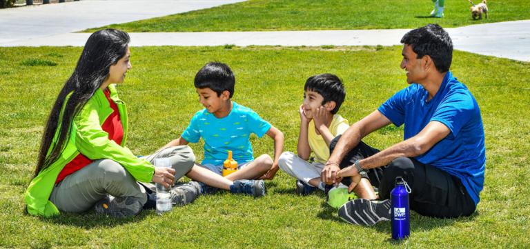Four Member family sitting on grass; from left to right, Mother, son, son and father smiling at each other sitting in half circle, with LADWP reusable water bottle to the father's feet