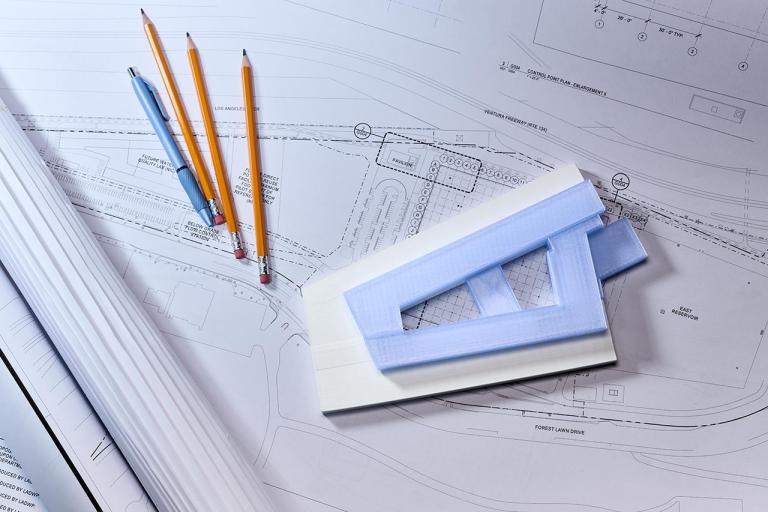 Drafting plans with three pencils, a pen and rule guide laying on top of the plans