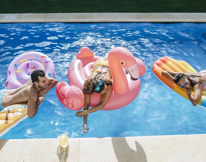 Three people laying on their stomachs on inflatables in a pool. The first inflatable is light pink with sprinkles and has a man laying on it. The second is a flamingo with a woman laying on it. The last is shaped like a red, yellow and blue popsicle with a woman laying on it, smiling and wearing sunglasses.