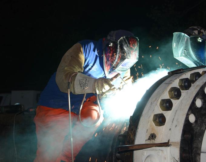 Two welders with welding masks on leaning over large pipe looking down as sparks arc from a welding torch.
