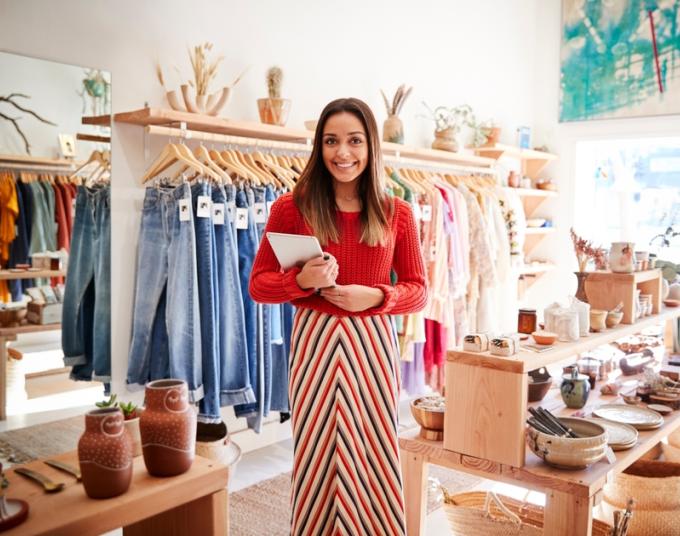 A smiling small business owner holds a tablet and stands in the middle of her retail store.