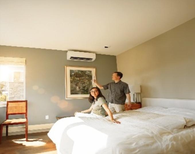 Man standing next to a woman seated on a bed, with a wall-mounted HVAC unit above a framed painting.